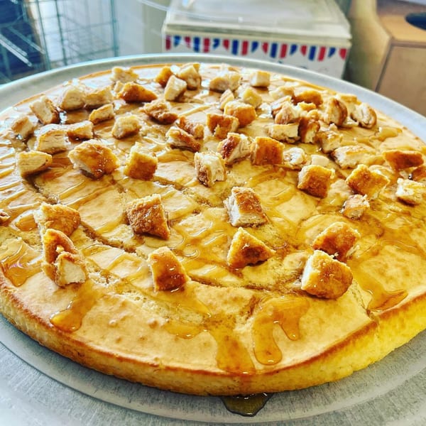 A golden waffle pizza topped with crispy chicken pieces and drizzled with syrup on a metal tray.