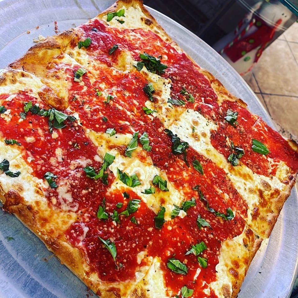 A square Luigi's Pizza with red sauce stripes, melted cheese, and fresh basil on a metal tray.