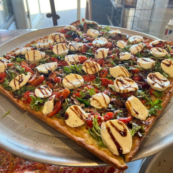 A square Luigi's Pizza with fresh mozzarella, tomatoes, greens, and balsamic glaze on a metal tray.